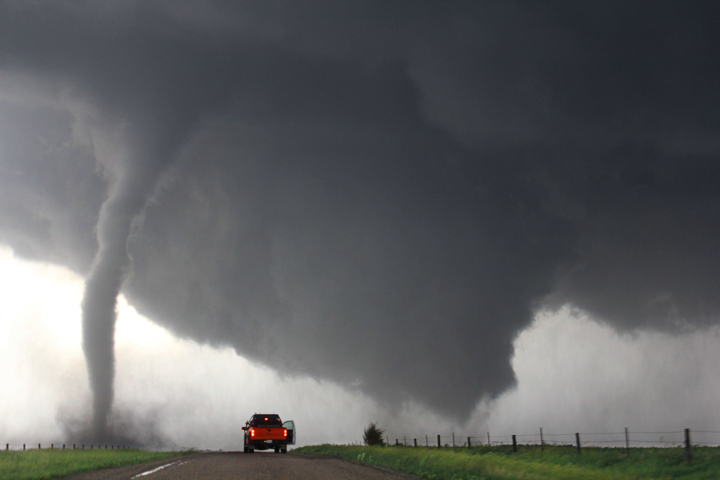 Een grote, dreigende tornado die een landweg nadert, met een oranje pick-up truck op de voorgrond.