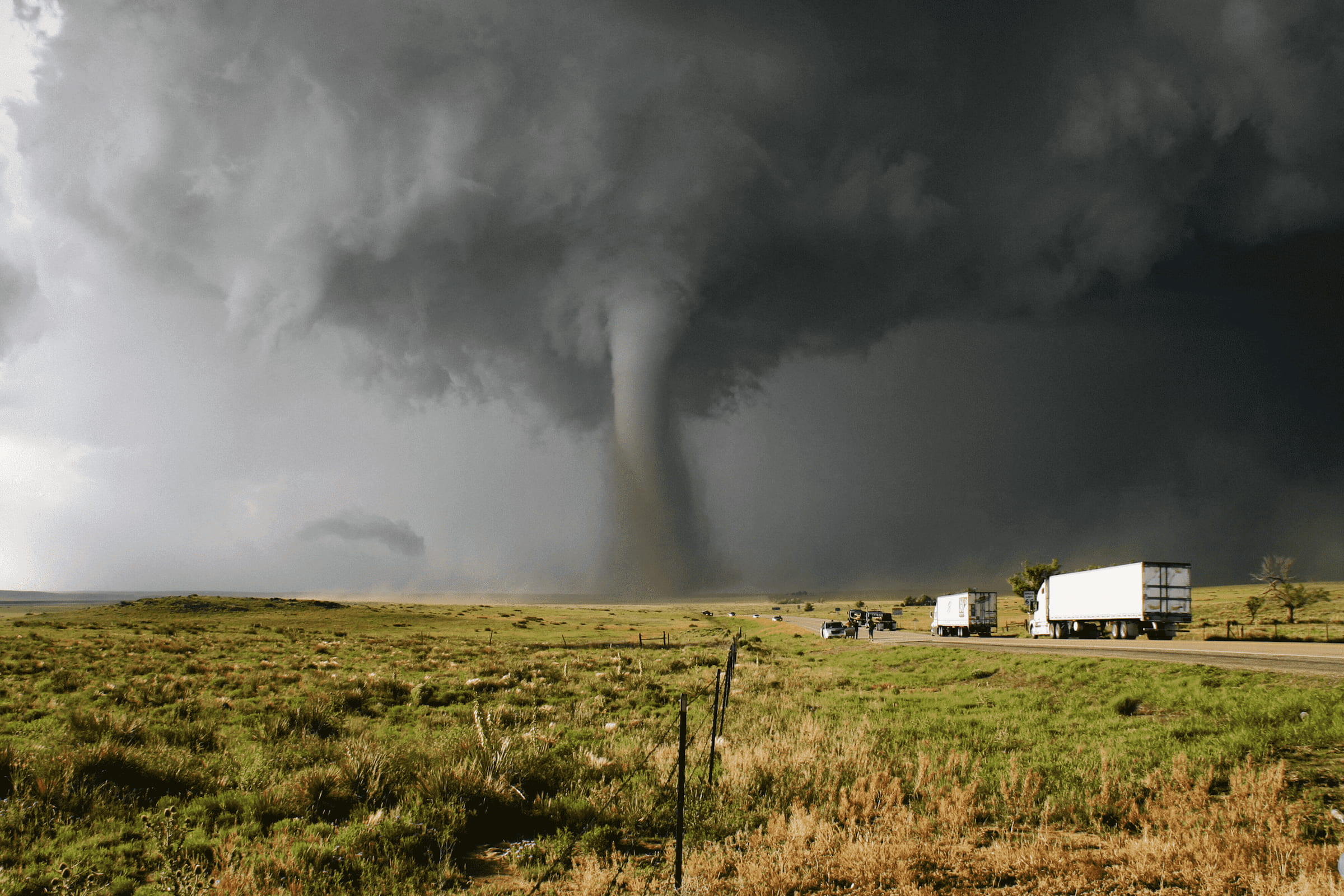Een grote, dreigende tornado die een landweg nadert, met vrachtwagens op de voorgrond.